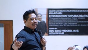 Seorang laki-laki berdiri memegang mic sedang menjelaskan materi pada audiens