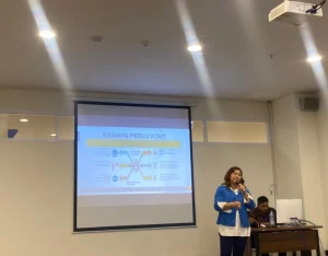 Dalam presentasi, seorang wanita menjelaskan informasi mengenai kawasan tanpa rokok di depan layar yang menampilkan grafik dan data