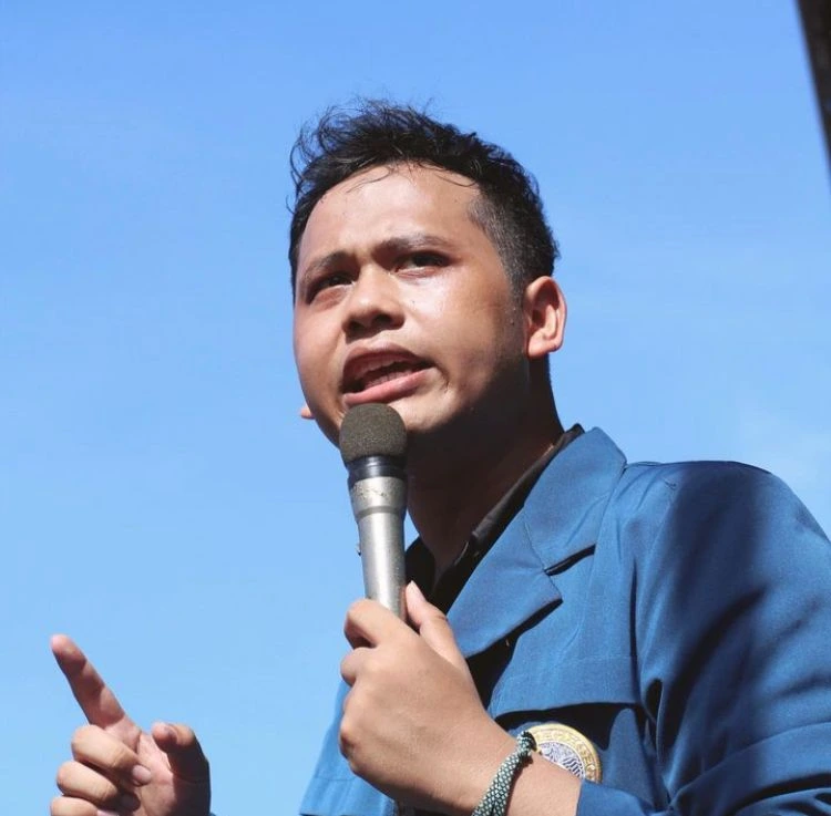 Seorang pria mengenakan jas almamater Unair berjaket biru sedang berbicara ke mikrofon.