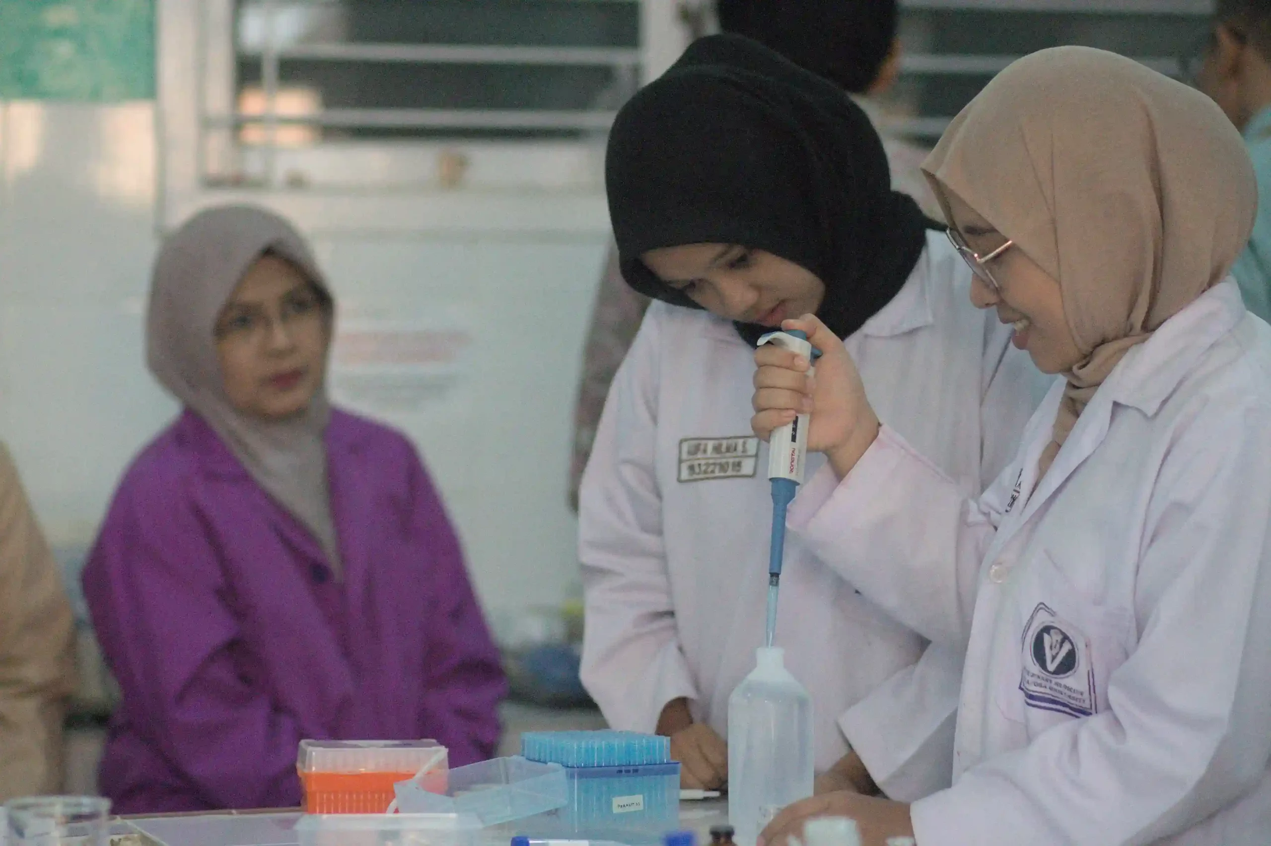 Sekelompok wanita mengenakan jas lab, berdiskusi di dalam laboratorium dengan peralatan ilmiah di sekitar mereka
