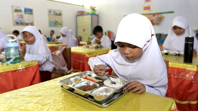 In a classroom setting, a young girl in a hijab enjoys a free nutritious meal.