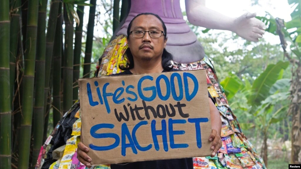 A man holds a sign reading "A Good Life Without Sachets" in front of a statue of a woman clutching plastic sachets.