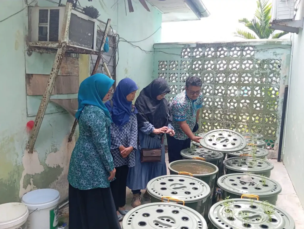 Three women and one man examine a bucket-based hydroponic system in a narrow residential space, part of a technology-driven community outreach initiative in Indonesia’s border region.