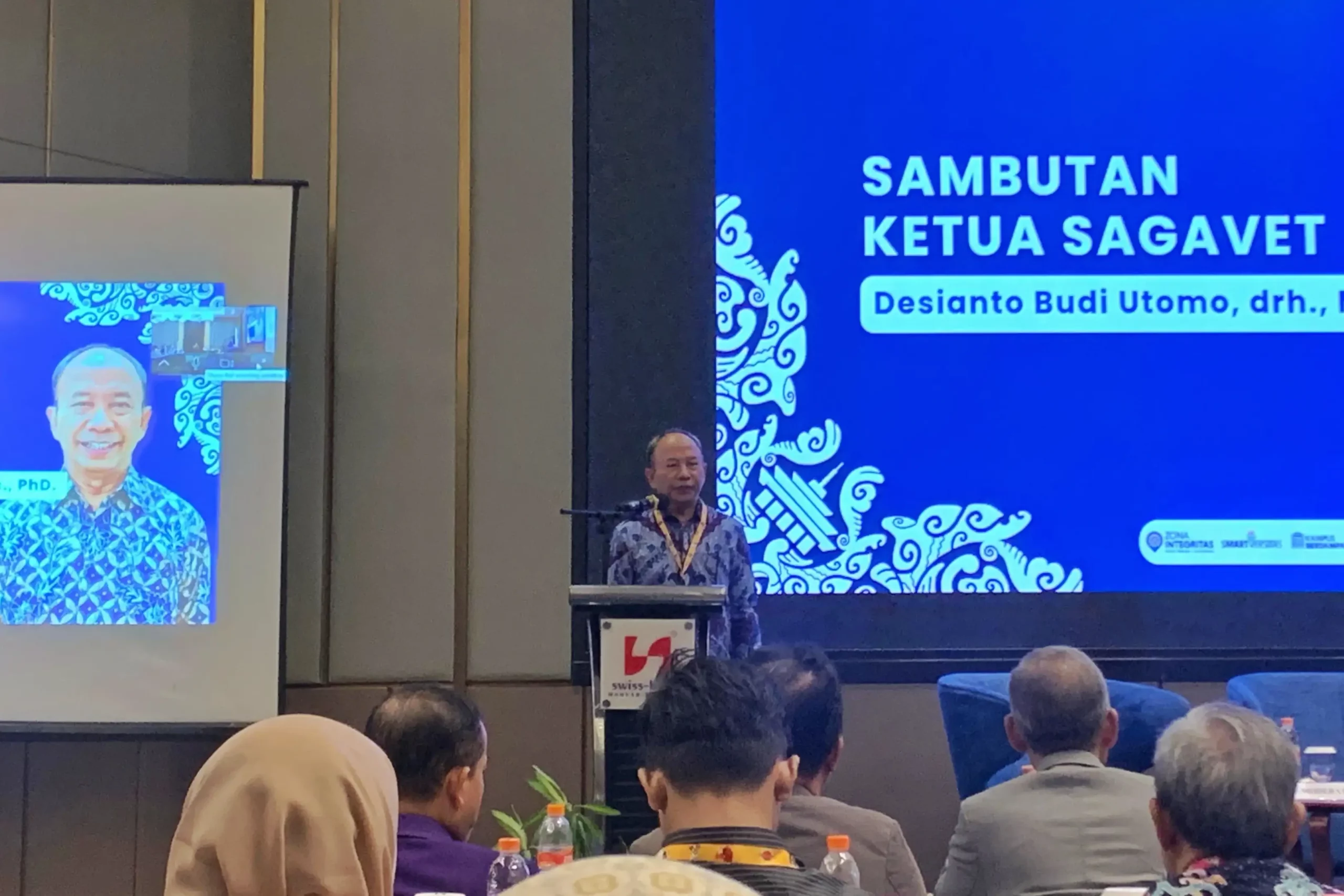 Seorang pria mengenakan baju batik berdiri di podium dengan latar belakang bertuliskan 'Sambutan Ketua SAGAVET Desianto Budi Utomo drh)