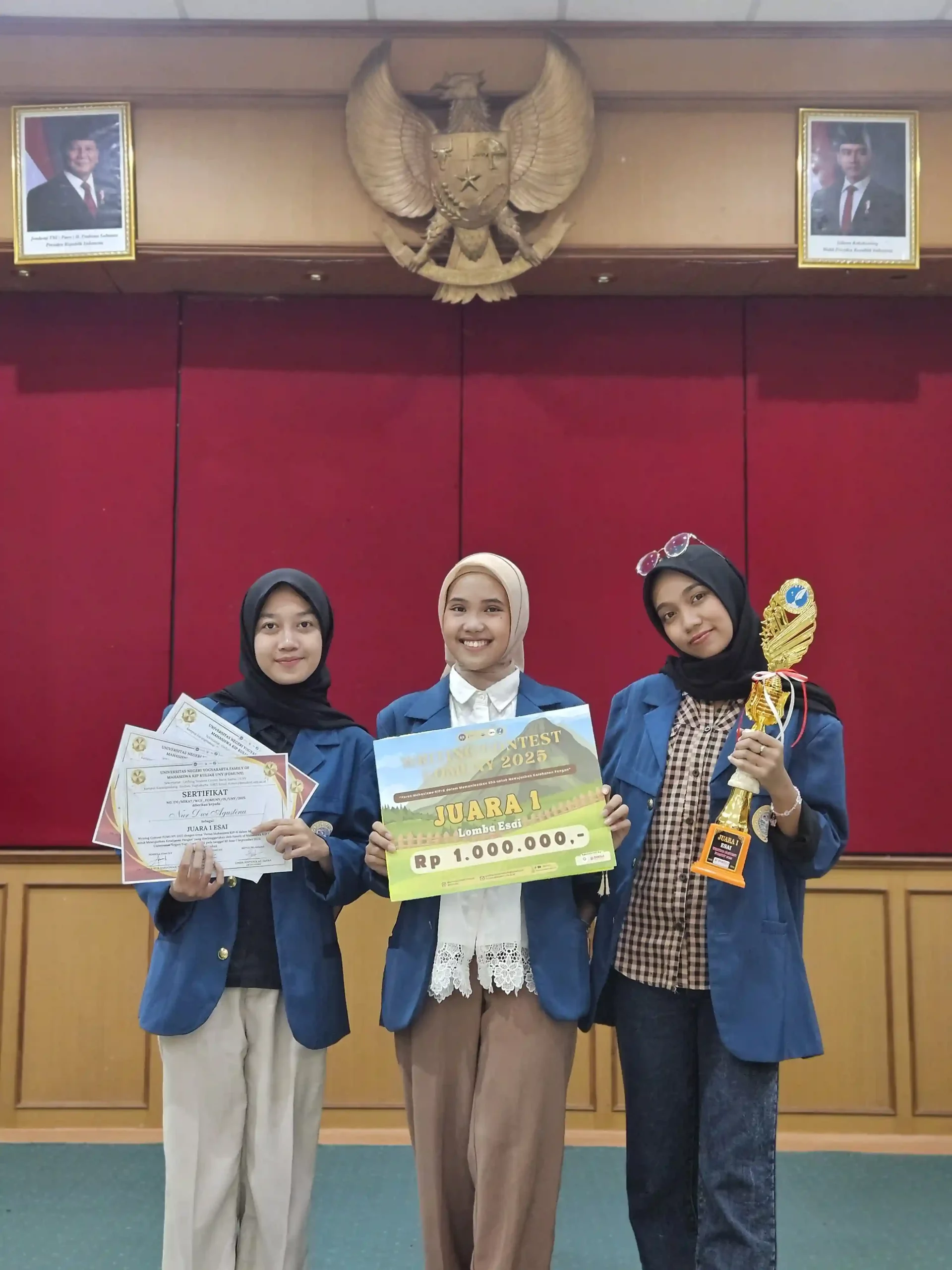Tiga orang perempuan mengenakan almamter berdiri membawa piala dan piagam penghargaan bertuliskan juara I lomba esai