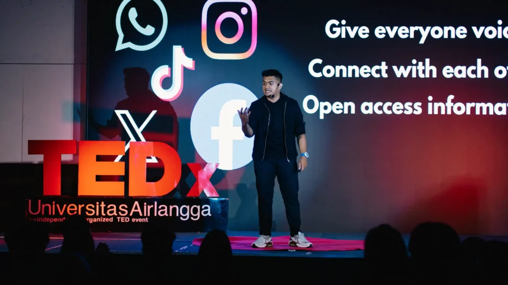 Seorang laki-lakimengenakan pakaian bernuansa hitam berdiri dengan background bertuliskan TEDxUniversitasAirlangga