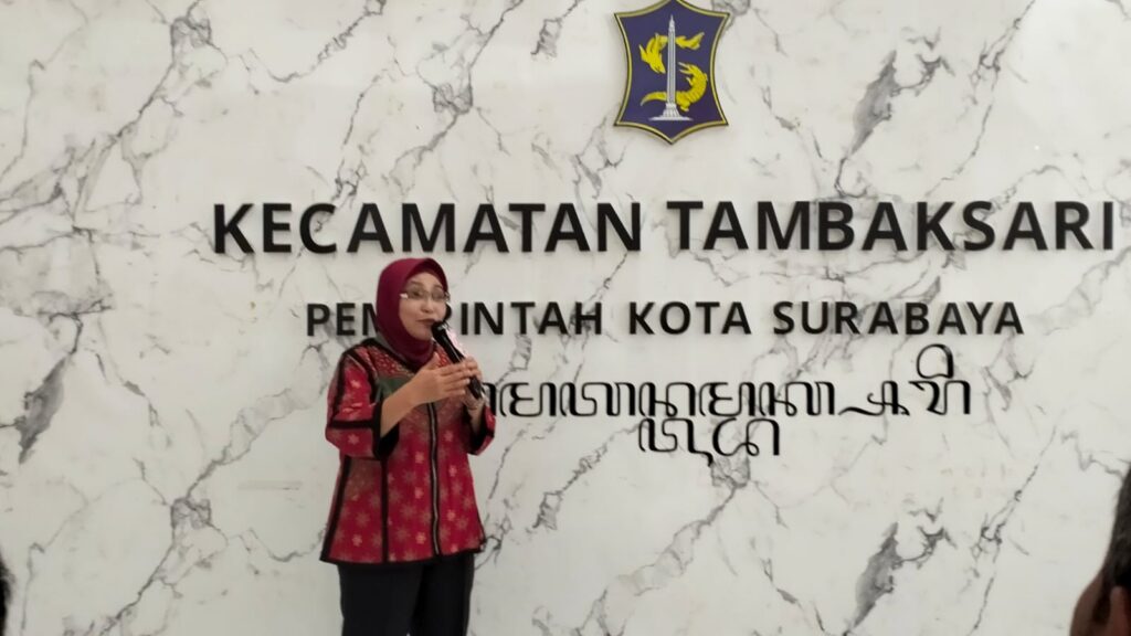 A woman wearing a hijab addresses an audience with a microphone in front of a marble-patterned backdrop labeled “Tambaksari Subdistrict, Surabaya City Government,” topped with the city’s blue and yellow emblem.