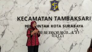 Seorang perempuan berhijab memegang mikrofon sambil berbicara di depan dinding bermotif marmer bertuliskan ‘Kecamatan Tambaksari Pemerintah Kota Surabaya’. Di atas tulisan terdapat lambang kota berwarna biru dan kuning