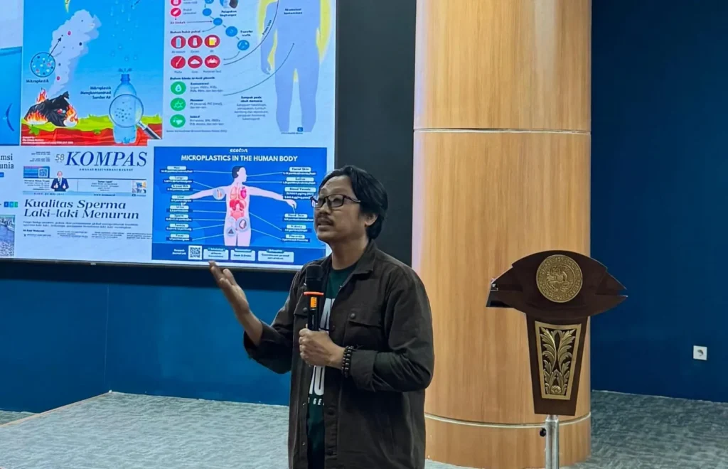 A male speaker holds a microphone while presenting in front of a large screen filled with graphs and infographics illustrating the risks of microplastics. He raises one hand mid-explanation, standing beside a wooden podium adorned with a university emblem.
