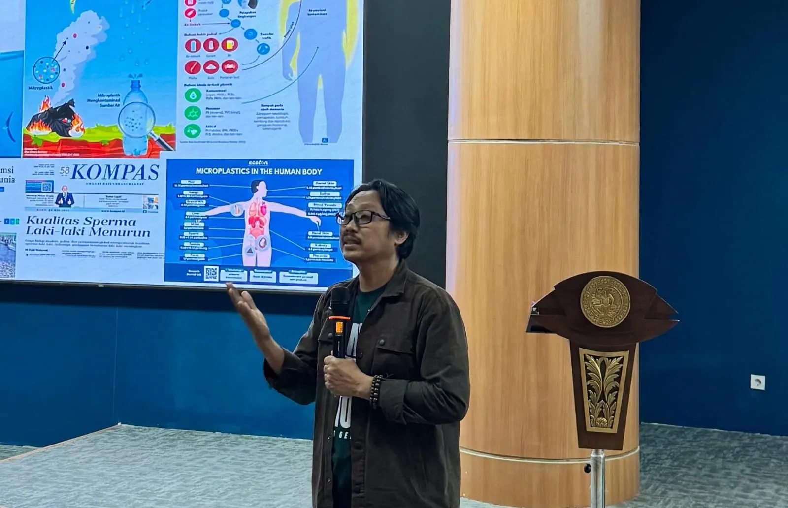 A male speaker holds a microphone while presenting in front of a large screen filled with graphs and infographics illustrating the risks of microplastics. He raises one hand mid-explanation, standing beside a wooden podium adorned with a university emblem.
