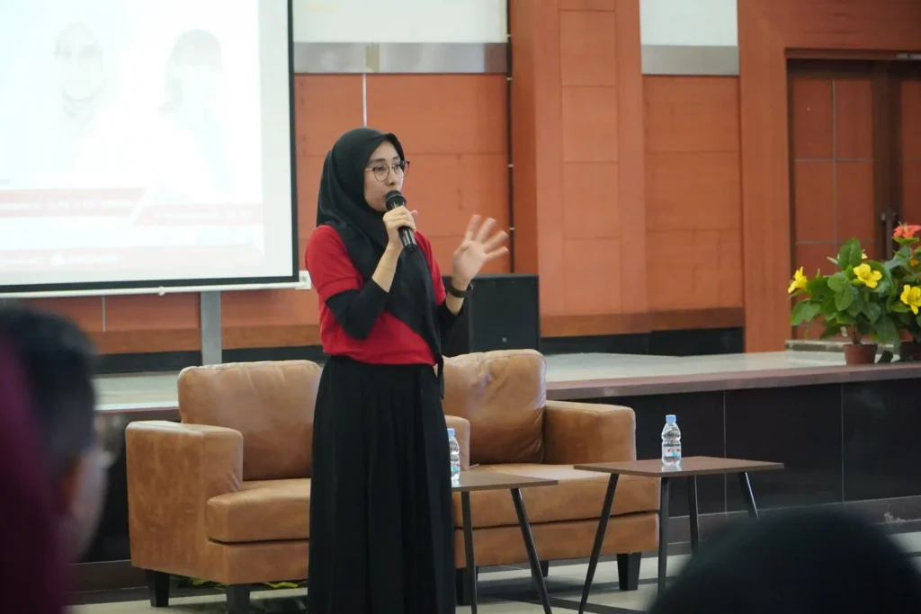 Seorang perempuan mengenakan baju merah dan bawahan hitam membawa mic berdiri menyampaikan materi pada audiens. Ia adalah Tri Pudy Asmarawati dr Sp PD K-PTI FINASIM menyampaikan materi pada seminar peringatan Hari HIV/AIDS yang digelar di Aula Dharmawangsa, Rumah Sakit UNAIR, Kampus MERR-C pada Sabtu (6/11/2025)