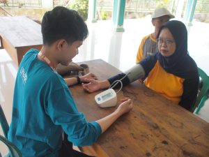Mahasiswa BBK 7 UNAIR sedang melakukan pemeriksaan tekanan darah pada peserta lansia menggunakan alat tensimeter sebagai bagian dari kegiatan skrining kesehatan dalam program BESTI di Balai Desa Barurejo. (Foto: Istimewa)