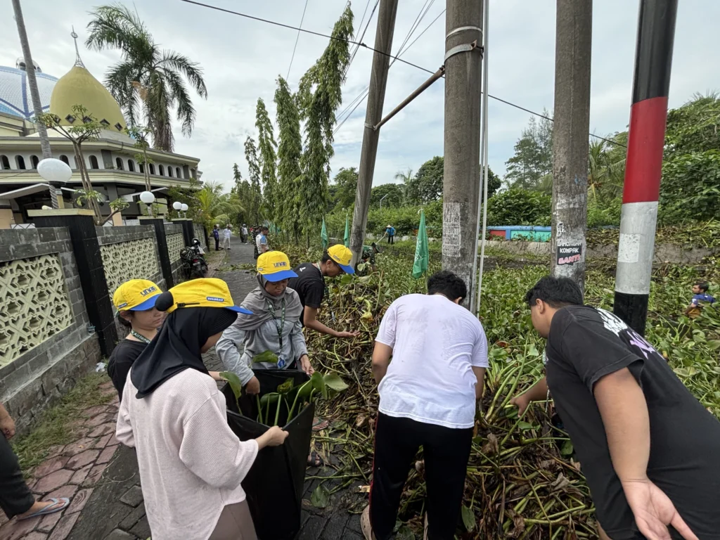 Mahasiswa UNAIR bergotong royong membersihkan tumpukan tanaman dan sampah di pinggir jalan dekat area pemukiman dan masjid