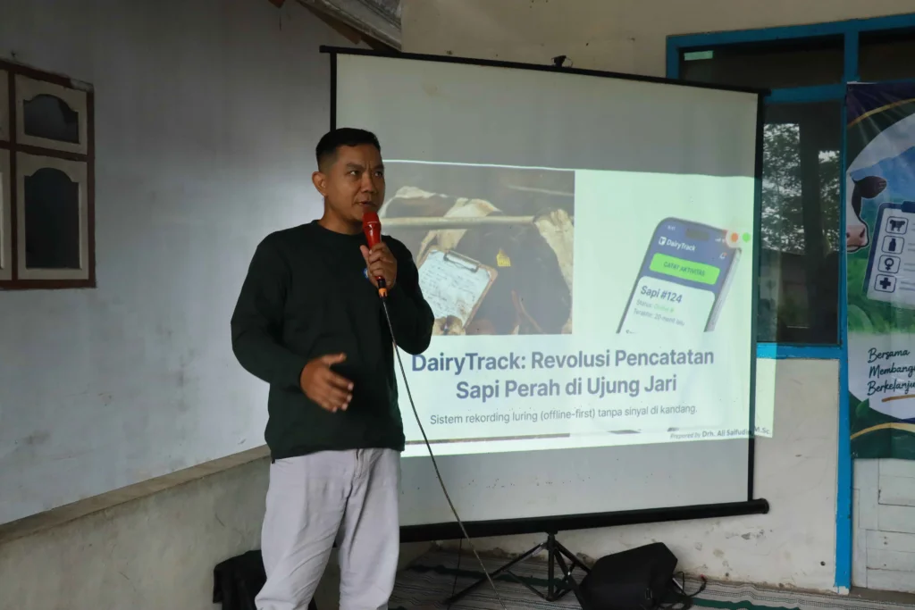 Seorang laki-laki mengenakan kaos berwarna hijau, berdiri dan memegang mikrofon, sedang berbicara di hadapan audiens. Ia adalah drh Ali Saifudin, anggota pengmas FKH UNAIR sedang mengenalkan aplikasi DairyTrack kepada poktan Wonosari, Desa Burno, Lumajang.